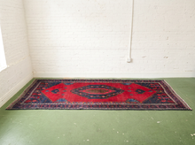 Load image into Gallery viewer, A rectangular vintage-style oriental rug with a red central field, dark navy blue corners, and intricate floral patterns, displayed on a green floor against a white brick wall.
