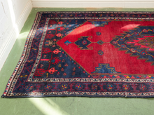 Load image into Gallery viewer, A traditional oriental area rug with a red central field, dark navy blue borders, and intricate multicolor patterns, laid out on a green floor.
