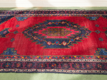 Load image into Gallery viewer, A traditional oriental area rug with a vibrant red field, a large central medallion in navy blue, and intricate geometric borders in cream, yellow, and green, laid out on a light green floor.
