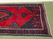 Load image into Gallery viewer, A traditional Persian-style area rug with a vibrant red central field, dark navy blue corners, and intricate multicolor geometric borders, laid out on a green floor.
