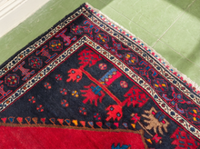 Load image into Gallery viewer, A close-up of an oriental area rug corner, showing a dark navy blue field with red, beige, and blue geometric motifs, bordered by red and cream bands, laid on a light green tiled floor.
