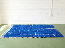 Load image into Gallery viewer, A rectangular blue shag rug with a white woven grid pattern and fringe on the left edge, laid out on a green floor against a white brick wall.
