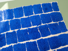 Load image into Gallery viewer, A close-up of a blue shaggy rug or mat with a white brick-patterned design, laid flat on a light green surface.

