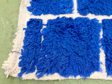 Load image into Gallery viewer, A close-up of a shaggy blue and white textured rug or textile laid on a green surface, showing thick looped fibers and a fringed edge.
