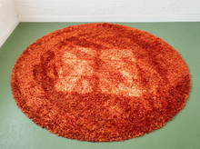 Load image into Gallery viewer, A round shag rug in rust orange with a lighter orange and cream patterned center, displayed on a green floor against a white brick wall.
