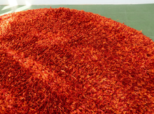 Load image into Gallery viewer, A close-up view of a round shag rug in a vibrant red-orange color, showing its thick, high-pile texture against a green floor background.
