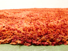 Load image into Gallery viewer, A close-up of a shaggy area rug with a textured, looped pile in warm shades of rust, orange, and red, shown on a green floor against a white wall.
