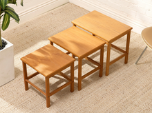 Load image into Gallery viewer, Three nested wooden nesting tables of varying heights in a warm light brown finish, arranged on a beige woven rug next to a potted plant.
