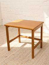 Load image into Gallery viewer, A square wooden side table or stool with a warm medium-brown finish, featuring a slatted front support and four straight legs, set on a woven rug against a white brick wall.
