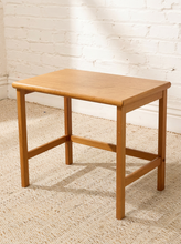 Load image into Gallery viewer, A small square wooden side table with a natural oak finish, featuring a simple rectangular top, four legs, and front stretchers, set on a textured beige rug against a white brick wall.
