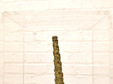 Load image into Gallery viewer, A close-up of a small cylindrical object with a textured, metallic gold-brown pattern, positioned vertically in front of a clear acrylic display block against a white brick wall background.
