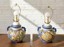 Load image into Gallery viewer, A pair of decorative ceramic table lamps with colorful hand-painted patterns, blue handles, and gold metal lamp frames, displayed on a wooden surface against a white brick wall.
