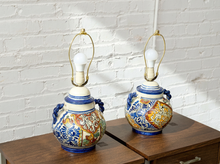 Load image into Gallery viewer, Two decorative ceramic table lamps with blue and gold handles, featuring colorful hand-painted floral and bird motifs, displayed on a wooden cabinet against a white brick wall.
