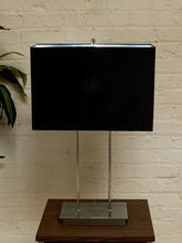 Load image into Gallery viewer, A modern table lamp with a large black rectangular shade supported by two slim vertical chrome legs and a square chrome base, placed on a wooden side table against a white brick wall.
