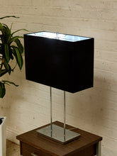 Load image into Gallery viewer, A modern table lamp with a matte black rectangular shade, dual polished silver metal legs, and a square silver base, shown lit on a wooden side table against a white brick wall.
