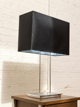 Load image into Gallery viewer, A modern table lamp with a large black rectangular fabric shade, a slim silver metal stem, and a square silver base, shown on a wooden side table against a white painted brick wall.
