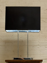 Load image into Gallery viewer, A modern table lamp with a large black rectangular shade supported by two slim vertical chrome legs and a circular chrome base, set against a light brick wall.
