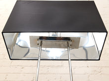 Load image into Gallery viewer, A rectangular black metal shade or panel with reflective silver mirrored inner walls and two chrome support legs, photographed against a white painted brick wall.

