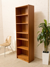 Load image into Gallery viewer, A tall, six-tier wooden bookcase with a warm medium-brown finish, positioned in a room corner against a white brick wall, flanked by a beige chair on the left and a potted green plant on the right.
