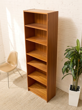 Load image into Gallery viewer, A tall, narrow wooden bookshelf with five open shelves, shown in a room setting against a white brick wall, next to a beige chair and a potted green plant.
