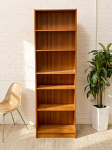 Load image into Gallery viewer, A tall, six-tier wooden bookcase with a warm medium-brown finish, positioned against a white brick wall in a styled room setting with a beige chair on the left and a potted green plant on the right.
