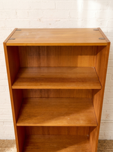 Load image into Gallery viewer, A mid-century modern wooden bookcase with three shelves, made of warm medium-brown wood with visible grain and small metal corner brackets, set against a white brick wall.
