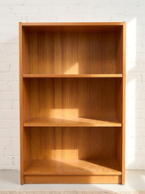 Load image into Gallery viewer, A three-tier wooden bookcase with a warm medium-brown finish, set against a white brick wall with soft sunlight casting shadows.
