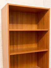 Load image into Gallery viewer, A three-tier wooden bookcase with a warm honey-wood finish, shown empty against a white painted brick wall.
