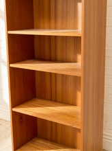 Load image into Gallery viewer, A tall wooden bookshelf with four open shelves, made of warm medium-brown wood with a visible grain pattern, set against a white brick wall.
