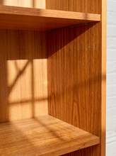 Load image into Gallery viewer, Close-up of a wooden bookshelf corner with warm sunlight casting shadows across the wood grain, next to a white brick wall.
