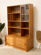 Load image into Gallery viewer, A mid-century modern wooden bookcase with a warm honey finish, featuring open shelves, two glass-front upper cabinets with round silver knobs, and two lower cabinet doors with small pull handles, set against a white brick wall.
