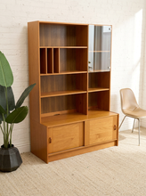 Load image into Gallery viewer, A mid-century modern wooden hutch with open shelves, a glass-paneled cabinet door, and two lower sliding doors, shown in a styled room setting with a brick wall, plant, and chair.
