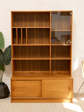 Load image into Gallery viewer, A mid-century modern wooden bookcase with a warm honey finish, featuring open shelves, a glass-front cabinet, and two lower storage doors, set against a white brick wall.
