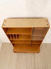 Load image into Gallery viewer, A mid-century modern wooden bookcase with a warm honey finish, featuring open shelves, a glass-front cabinet, and a pull-out bottom drawer, set against a white brick wall and light beige rug.
