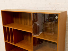 Load image into Gallery viewer, A mid-century modern wooden bookcase with a warm honey finish, featuring open shelving, a slatted side panel, and a glass-front cabinet section on a white brick wall background.
