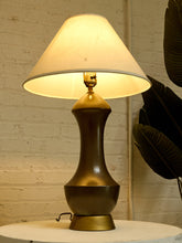 Load image into Gallery viewer, A vintage-style table lamp with a warm white conical shade and a dark metallic bronze-colored base, shown lit on a white table against a white brick wall with a plant leaf in the background.
