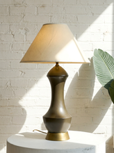 Load image into Gallery viewer, A table lamp with a tapered cream fabric shade and a metallic bronze-finished ceramic base, styled on a white pedestal against a white brick wall with sunlight and shadows.
