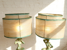Load image into Gallery viewer, A pair of vintage table lamps with tiered beige fabric shades featuring teal-green stitched trim and draped over sculptural green metal bases, set against a white brick wall with warm sunlight and shadows.
