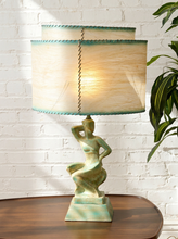 Load image into Gallery viewer, A table lamp with a sculpted green female figure base and a two-tiered beige fabric shade with teal trim, shown on a wooden table against a white brick wall with a plant nearby.
