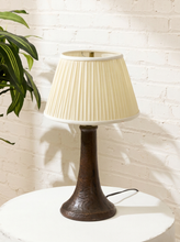 Load image into Gallery viewer, A table lamp with a pleated cream shade and a dark brown textured base, sitting on a white round table against a white brick wall with a green plant leaf on the left.

