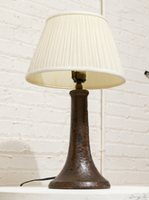Load image into Gallery viewer, A table lamp with a textured, hammered dark brown metal base and a pleated off-white fabric shade, set against a white brick wall background.
