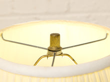 Load image into Gallery viewer, Close-up of a vintage table lamp shade with a cream-colored fabric body, a brass central finial, and a metal spider support structure inside a lit shade.
