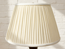 Load image into Gallery viewer, A cream-colored pleated fabric lampshade with white trim, shown on a table against a white painted brick wall background.
