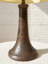 Load image into Gallery viewer, A rustic hammered metal table lamp base in a dark bronze finish, shown on a textured white surface against a white brick wall background.
