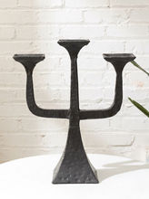 Load image into Gallery viewer, A black metal three-arm candelabra with a textured, hammered finish, displayed on a white surface against a white brick wall background.
