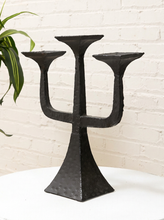 Load image into Gallery viewer, A black metal three-arm candelabra with a textured, hammered finish, displayed on a white table against a white brick wall background.

