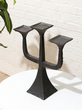 Load image into Gallery viewer, A black cast iron three-arm candelabra with a textured, hammered finish, displayed on a white round table against a white brick wall background.
