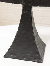Load image into Gallery viewer, A close-up of a black textured metal sculpture or stand with a hammered, dotted surface finish, resting on a white surface against a light brick wall background.
