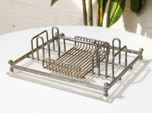 Load image into Gallery viewer, A vintage silver metal washstand or dish rack with a slatted center tray, set on a white tablecloth with a white brick wall and green plants in the background.
