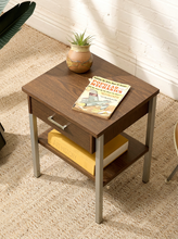 Load image into Gallery viewer, A square wooden end table with a single drawer and open lower shelf, styled with a small potted plant and a magazine on top, set on a textured beige rug against a white brick wall.
